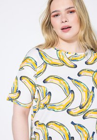 Camiseta de manga corta con una base blanca y un estampado colorido de plátanos en amarillo, azul y rosa. Cuello redondo, ajuste relajado.