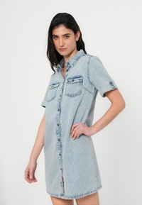 Φόρεμα πουκάμισο από τζιν σε ανοιχτό μπλε, με κοντά μανίκια, κουμπιά μπροστά, δύο τσέπες στο στήθος και ανοίγματα στα πλάγια στο τελείωμα.
