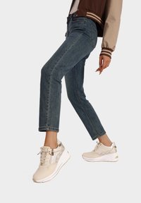 Sneakers atletici beige con dettagli dorati lucidi e perforazioni, abbinati a jeans blu aderenti e una giacca bomber marrone.