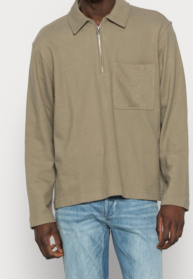 Homme portant un polo à manches longues beige avec une fermeture éclair à l'avant et une poche poitrine, associé à un jean en denim bleu clair.
