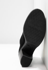 Zapato de ante negro con suela de goma texturizada, que presenta un tacón delgado y un diseño sencillo. El zapato está colocado sobre un fondo blanco.