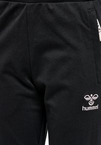Schwarze Sporthose mit glatter Textur, ausgestattet mit einem weißen gestickten Logo an der Seite. Enthält einen elastischen Bund und Seitentaschen.