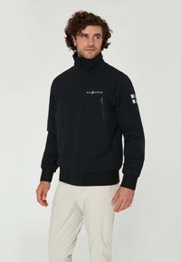 Svart softshell-jacka med hög krage, framdragkedja och två dragkedjefickor på bröstet. Har logotyp på vänster sida och elastiska ärmslut.