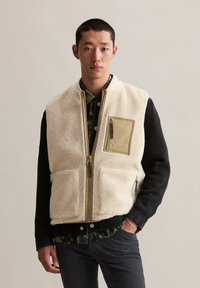 Jonge man in een beige fleece vest met vovectoren over een zwarte jas en een groen patroonhemd, staande met één hand in zijn zak.