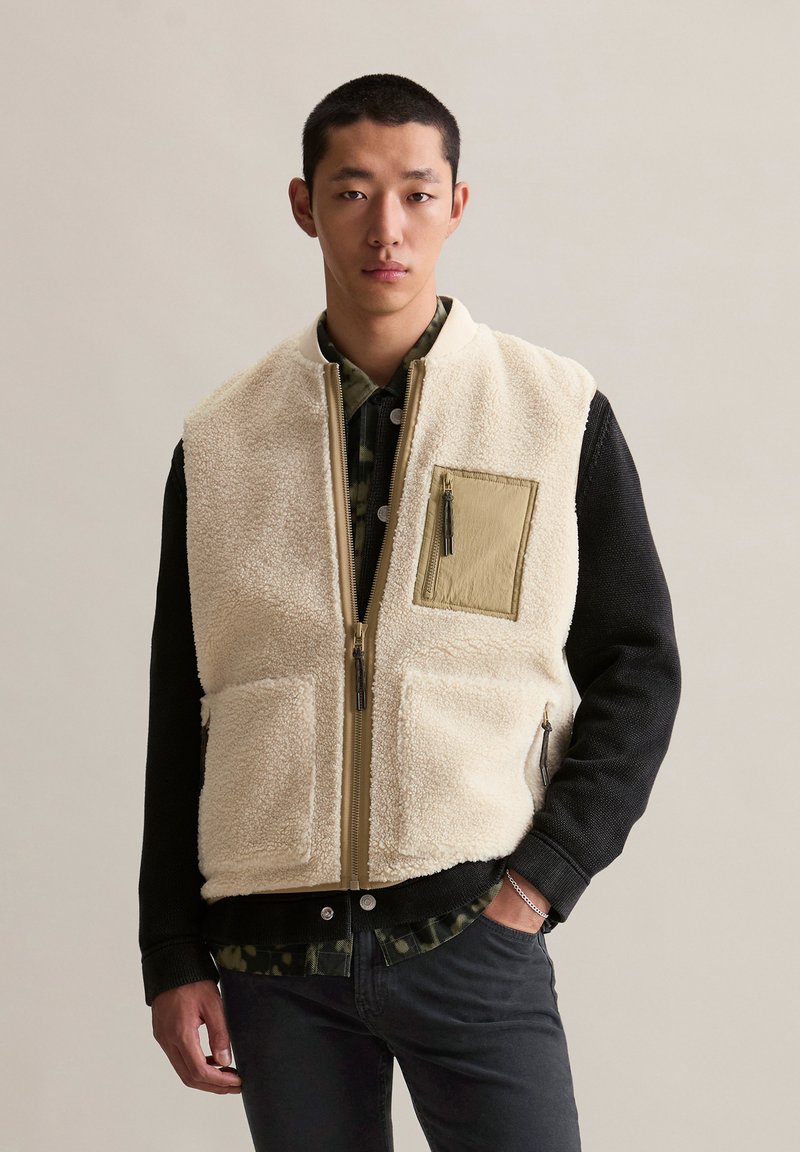 Jonge man in een beige fleece vest met vovectoren over een zwarte jas en een groen patroonhemd, staande met één hand in zijn zak.