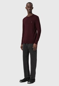 Maglione a maglia bordeaux con maniche lunghe, scollatura rotonda e piccolo dettaglio del logo, abbinato a jeans grigio scuro e stivali neri con accenti argentati.