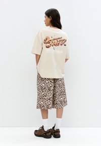 Tricou oversized bej cu grafică tematică despre biscuiți și text pe spate, asortat cu pantaloni scurți cu imprimeu leopard maro și adidași negri.