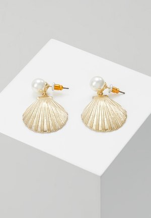 Gouden oorbellen in schelpvorm met ronde witte parelstekers en gouden pennen, tentoongesteld op een wit voetstuk tegen een grijze achtergrond.