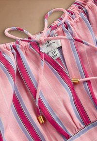 Rozā, sarkanas un lavandas krāsas svītrains audums ar savilktu elastīgu kakla daļu un sasietu auklu ar zelta metāla galiņiem un apģērba izmēra etiķeti.