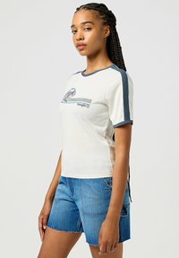 Camiseta blanca con mangas cortas azules, que presenta un gráfico de ola y el texto "Wrangler". Combinada con pantalones cortos de mezclilla azules. Ajuste y diseño casual.