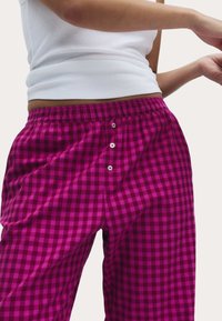 Różowo-czarne spodnie piżamowe w kratę z elastycznym paskiem i trzema guzikami z przodu. Tkanina wydaje się miękka i przewiewna.