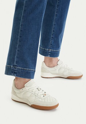 Zapatillas blancas acolchadas con suelas de goma, usadas con vaqueros azules, mostrando las piernas bajas y los tobillos contra un fondo claro y liso.