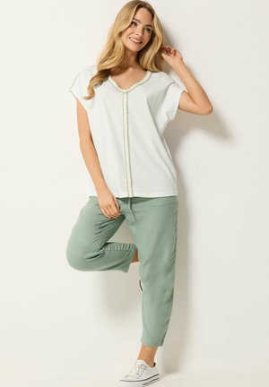 Smilende kvinde med langt blondt hår iført en hvid V-hals top og lysegrønne bukser, løfter det ene ben og står mod en ensfarvet baggrund.