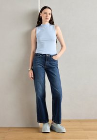 Lichtblauw mouwloos topje, hoge hals, gecombineerd met donkerblauwe wijd-uitlopende jeans en grijze instapschoenen, met soepele texturen en een eenvoudig ontwerp.