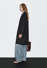 Czarny, oversize płaszcz o prostym kroju, połączony z jasnoniebieskimi, szerokimi dżinsami i sandałami. Zawiera brązową torbę i kilka pierścionków.