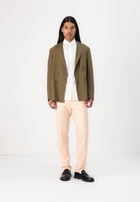Olijfgroen dubbelzijdig blazer over een wit shirt, gecombineerd met lichtroze broeken en zwarte schoenen, gepresenteerd tegen een witte achtergrond.