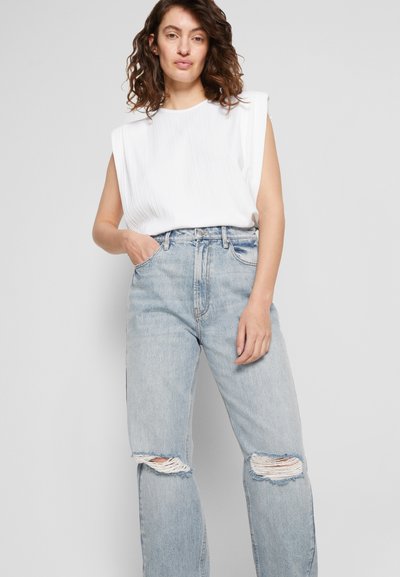 Top branco sem mangas com textura plissada, combinado com jeans de cintura alta de tom azul claro apresentando rasgos nos joelhos.