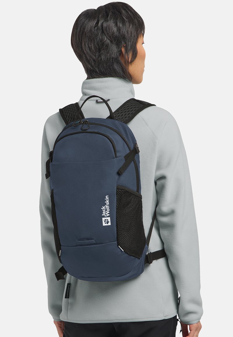 Persona che indossa una giacca in pile grigia con uno zaino Jack Wolfskin blu navy, dotato di spallacci imbottiti neri e tasche laterali in rete.