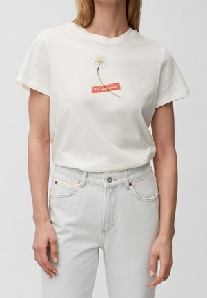 Frau trägt weißes T-Shirt mit Gänseblümchen und rotem Schriftzug "let love grow", kombiniert mit hellblauen Jeans mit hoher Taille vor einfachem Hintergrund.