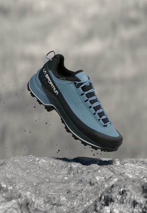 Zapato de senderismo La Sportiva azul y negro en el aire sobre terreno rocoso con partículas de tierra, con Gore-Tex y suela de dibujo agresivo.