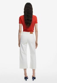 Weiße Schlagjeans mit hoher Taille, hinten mit einem Lederpatch versehen, kombiniert mit einem figurbetonten roten T-Shirt und blauen Absatzsandalen.