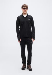 Μαύρο τοπ με φερμουάρ στο ένα τέταρτο και μακρυμάνικο, μαζί με αντίστοιχο παντελόνι, φορεμένα με καφέ παπούτσια πεζοπορίας. Το ύφασμα φαίνεται ελαφρύ και ελαστικό.