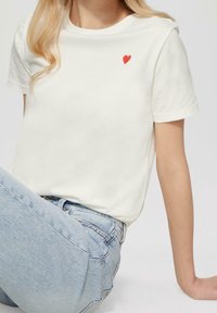 Hvid bomulds T-shirt med korte ærmer, der har et lille rødt hjerte-design på venstre bryst. Stylet med lyseblå denim jeans.