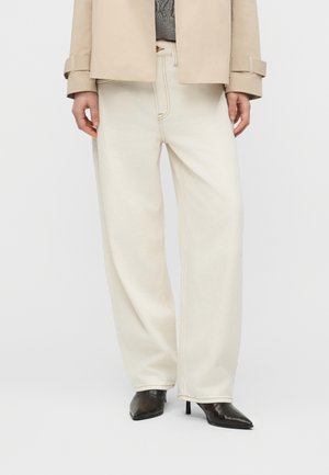 Γυναίκα με εκρού παντελόνι φαρδύ, μπεζ σακάκι, γκρι πουλόβερ και μαύρα μυτερά μπότες με τακούνι, στέκεται μπροστά σε λευκό φόντο.