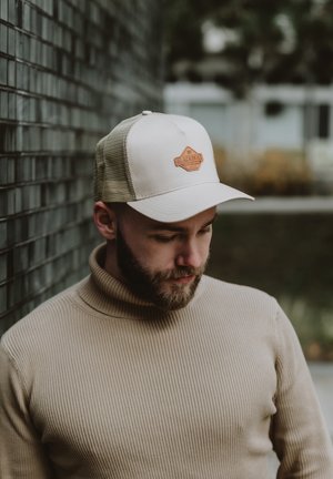 Beige trucker cap with a brown leather patch. Ribbed beige turtleneck sweater. Typical mesh back. Subject looking down against a dark wall.