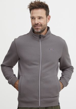 Grå zip-genser med høy krage, ribbede mansjetter, sidelommer og en liten logo detalj på brystet. Glatt teksturert stoff.