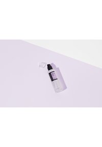 Bouteille en plastique transparent avec un bouchon pompe noir, ornée d'une étiquette violette et d'un capuchon transparent, placée sur une surface violette claire.