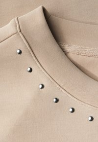Maglietta beige con scollatura rotonda decorata con accenti di borchie metalliche disposte in linea, realizzata in un tessuto morbido e liscio.
