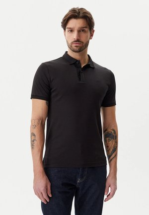 Polo shirt nera a maniche corte con colletto e placca a bottoni; realizzata in tessuto liscio; indossata con jeans in denim blu scuro.