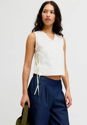 Kobieta w białym, bezrękawnym topie typu wrap z wiązaniami po bokach i luźnych ciemnoniebieskich spodniach, trzymająca pasek oliwkowej torby.