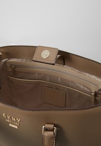 Sac à main en cuir fauve avec ferrures dorées. L'intérieur comprend une poche zippée et plusieurs compartiments, marqués du logo DKNY.