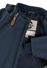 Laivastonsininen takki, jossa sileä ja vedenpitävä ulkokerros. Varustettu resorihihoilla, vetoketjullisella edellä ja nahkaisella vetoketjun vetimellä. Sisäosassa etiketti.