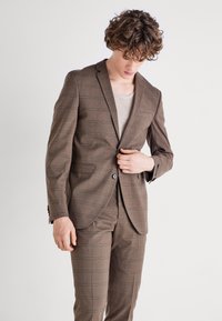 Costum cu carouri maro, format din sacou cu două butoane și pantaloni asortați. Țesătura are o textură netedă, cu detalii subtile ale modelului și cusături contrastante.