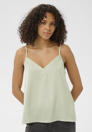 Femme aux cheveux bouclés portant un haut vert clair sans manches à col en V et un pantalon foncé, debout les mains derrière le dos.