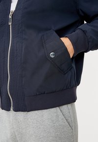 Hand in de zak van een marineblauwe ritsjack met drukknoopflap, gecombineerd met lichtgrijze broek tegen een eenvoudige witte achtergrond.