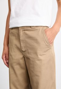 Pantaloni in cotone beige con taglio dritto, dotati di tasca frontale e passanti per cintura. Il tessuto ha una texture liscia e resistente.