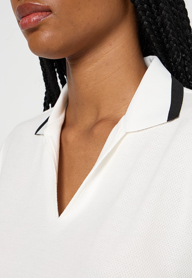 Polo blanc en tissu texturé, avec un col en V et des finitions noires contrastantes sur le col. Vue en gros plan avec des détails minimaux.