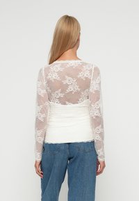 Biela čipkovaná blúzka s dlhými rukávmi, zdobená kvetinovými vzormi. Hrudník je zberaný s textúrovanou, pevnú bielou podšívkou. Nosené s džínsami.