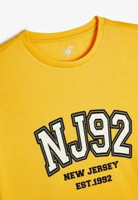 Keltainen urheilupaita, joka on valmistettu teksturoidusta kankaasta. Ominaisuuksiin kuuluu musta ja valkoinen printti "NJ92" ja sen alapuolella "NEW JERSEY EST.1992". Pyöreä kaula-aukko.
