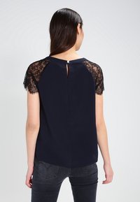 Haut bleu marine avec des manches courtes en dentelle, comportant un dos en trou de serrure et des détails en boutons. Le tissu lisse et le motif délicat de dentelle rehaussent le design.