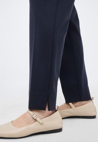 Κάτω άκρα ντυμένα με μπλε σκούρα παντελόνια που στενεύουν προς τον αστράγαλο με σχισμές στα πόδια, συνδυασμένα με μπεζ μπαλαρίνες Mary Jane με λουράκια και αγκράφες.