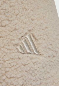 Textura suave de color crema con un logo de Adidas en plateado metálico sutilmente en relieve, en forma de tres rayas.