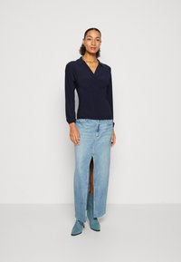 Marineblå langermet topp med omslagsdesign, parret med en lys blå denimskjørt med frontslitt og slitt kant, komplettert med teal støvler.