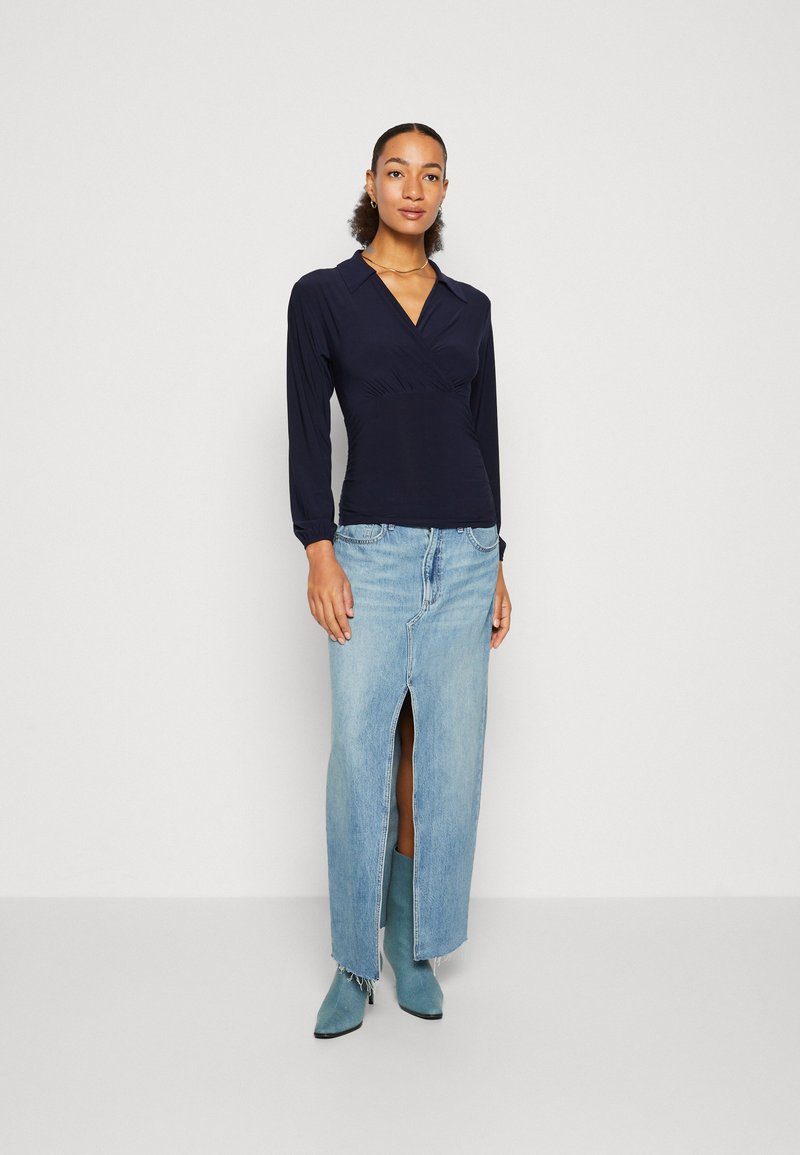 Marinefarbener Langarmtop mit Wickeldesign, kombiniert mit einem hellblauen Denimrock mit vorderem Schlitz und fransigem Saum, ergänzt durch türkisfarbene Stiefel.