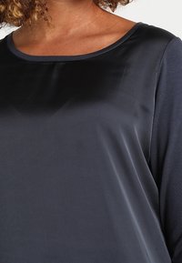 Frau trägt ein lange Ärmel-Oberteil aus dunkelgrauem Satin mit rundem Ausschnitt und strukturierten Ärmeln.