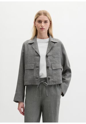 Woman wearing a grey buttoned jacket with front pockets over a white shirt, paired with matching grey drawstring pants, standing against a plain background.